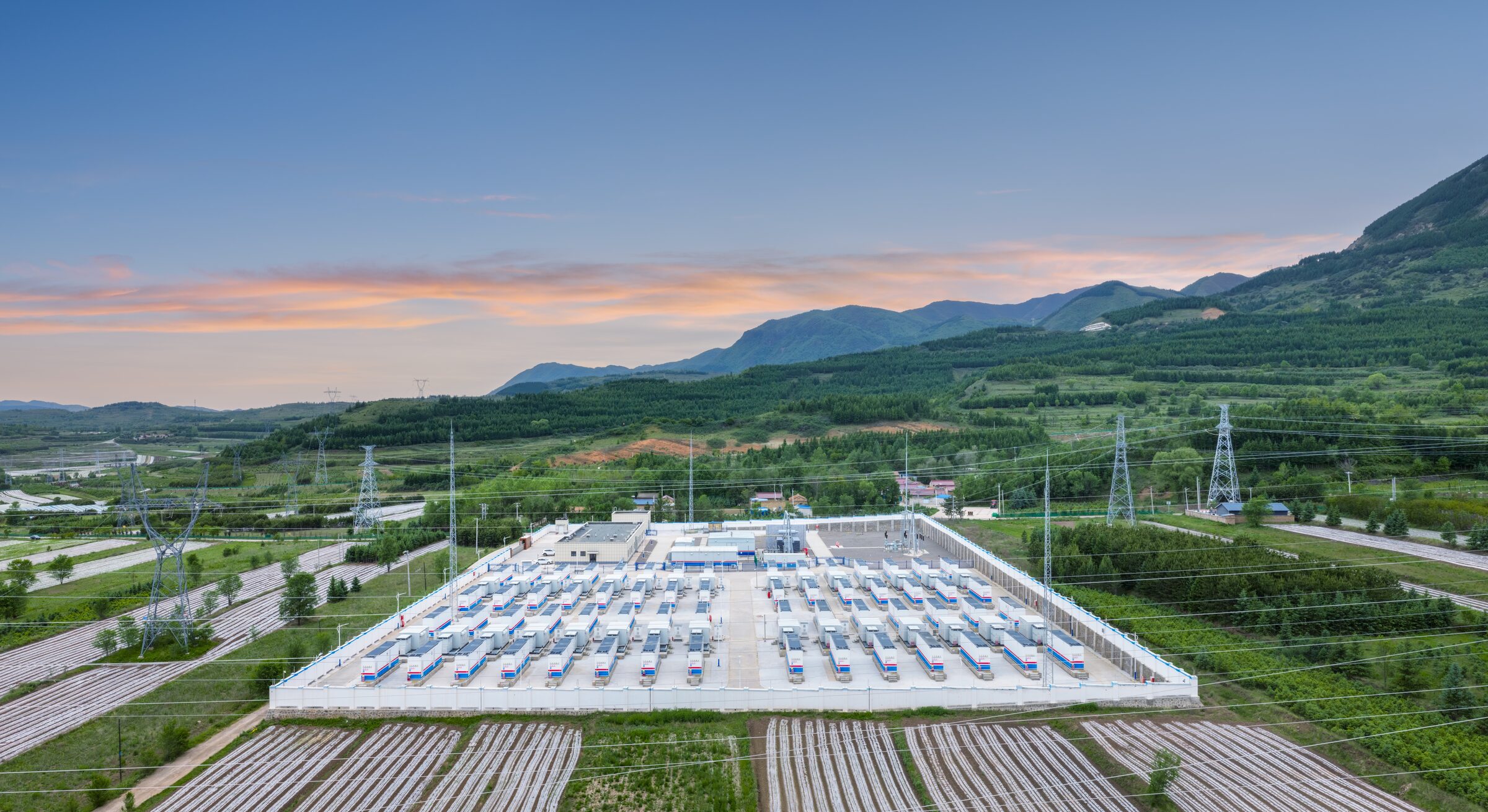 Aerial view at dusk of a large battery storage site on a valley floor, with mountains and farmland behind it